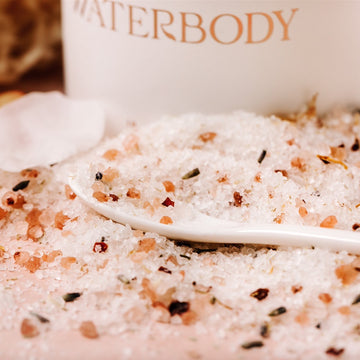 Sea salts and Himalayan pink salts flecked with bits of flower petals are spilled out in front of a white paperboard tube package